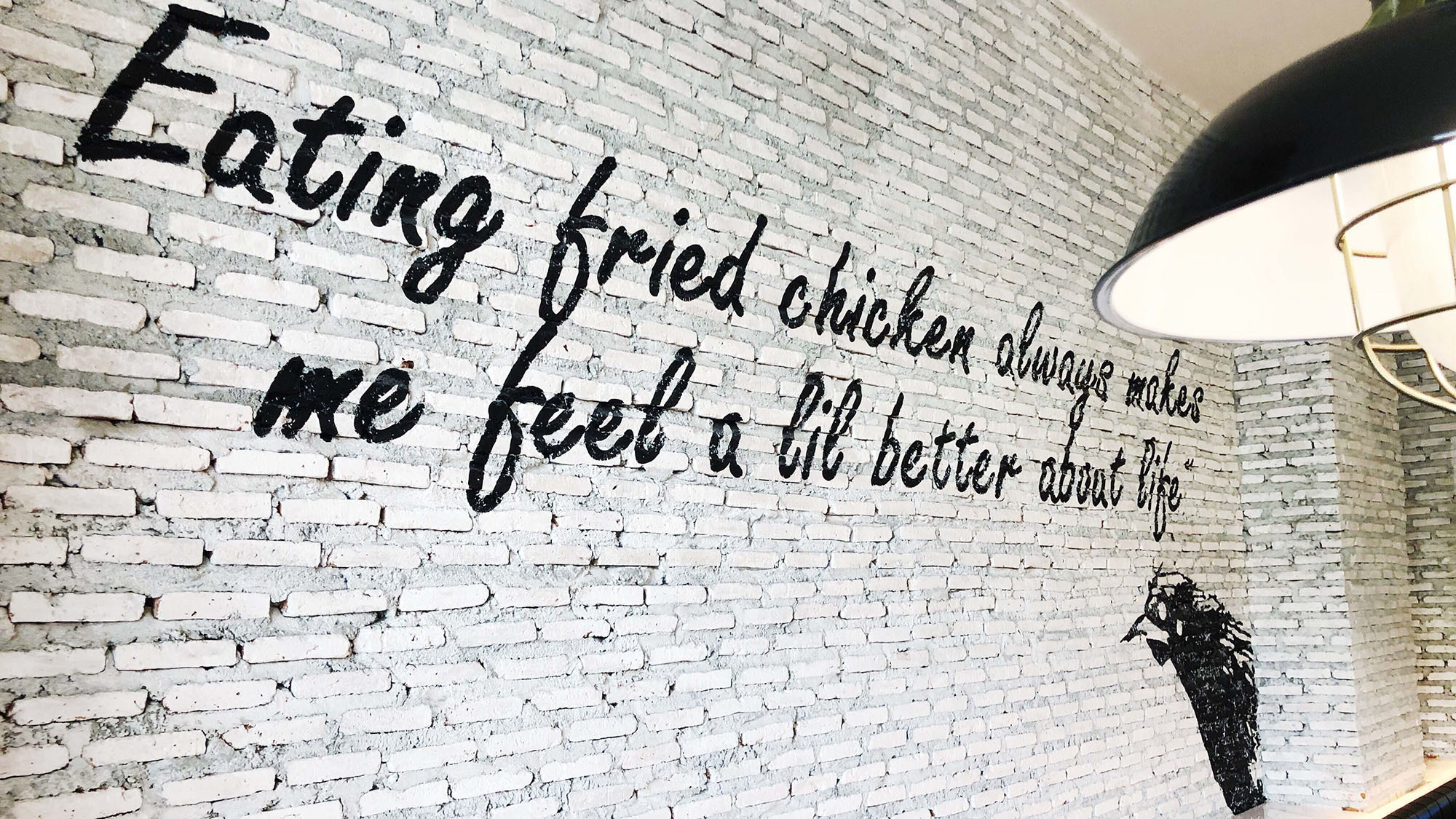 Weiße Ziegelwand mit schwarzer Handlettering-Zitat über frittierten Hühnchen, Pendelleuchte und Hühnersilhouette – kreative Designelemente für Systemgastronomie-Employer Branding oder Markenstrategien.