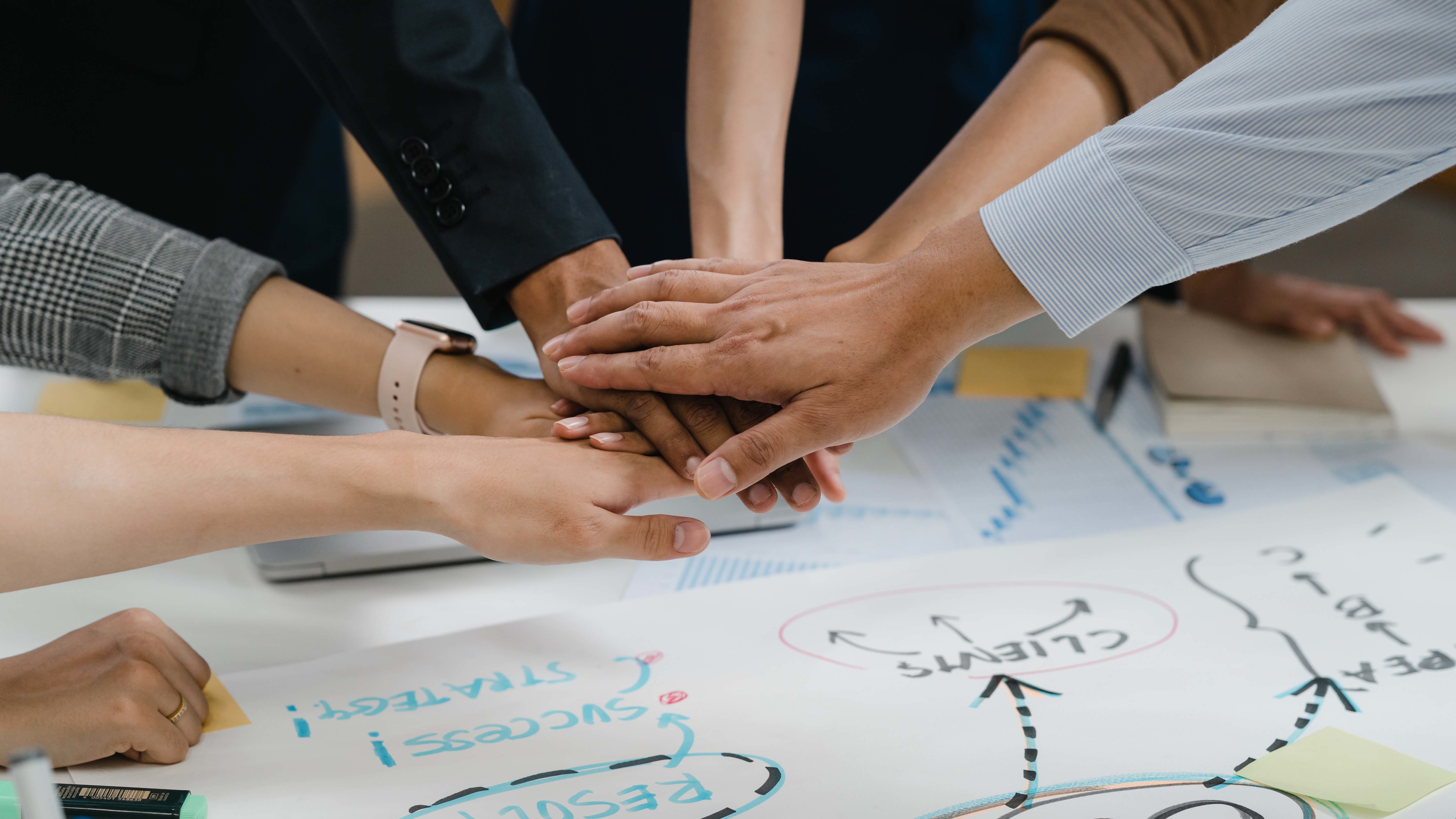 Team von N3W – creative strategies demonstriert mit gestapelten Händen Zusammenarbeit und gemeinsame kreative Strategieentwicklung an einem Tisch mit Diagrammen und Notizen.