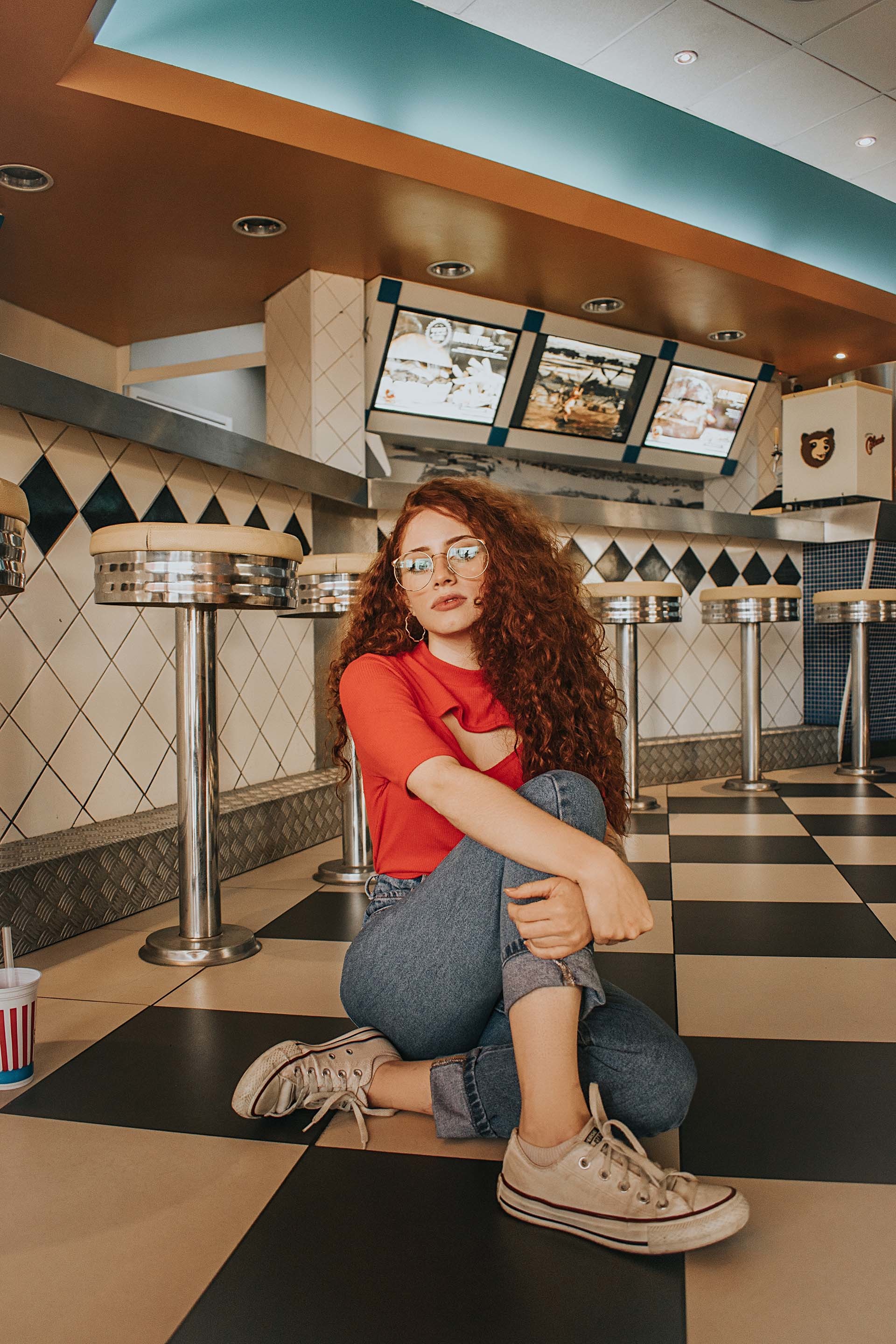 Frau mit roten Locken sitzt auf dem Boden eines retro-stilisierten Diners mit Schachbrettmuster – Bild für Employer Branding in der Systemgastronomie, entwickelt von der Kreativagentur N3W.