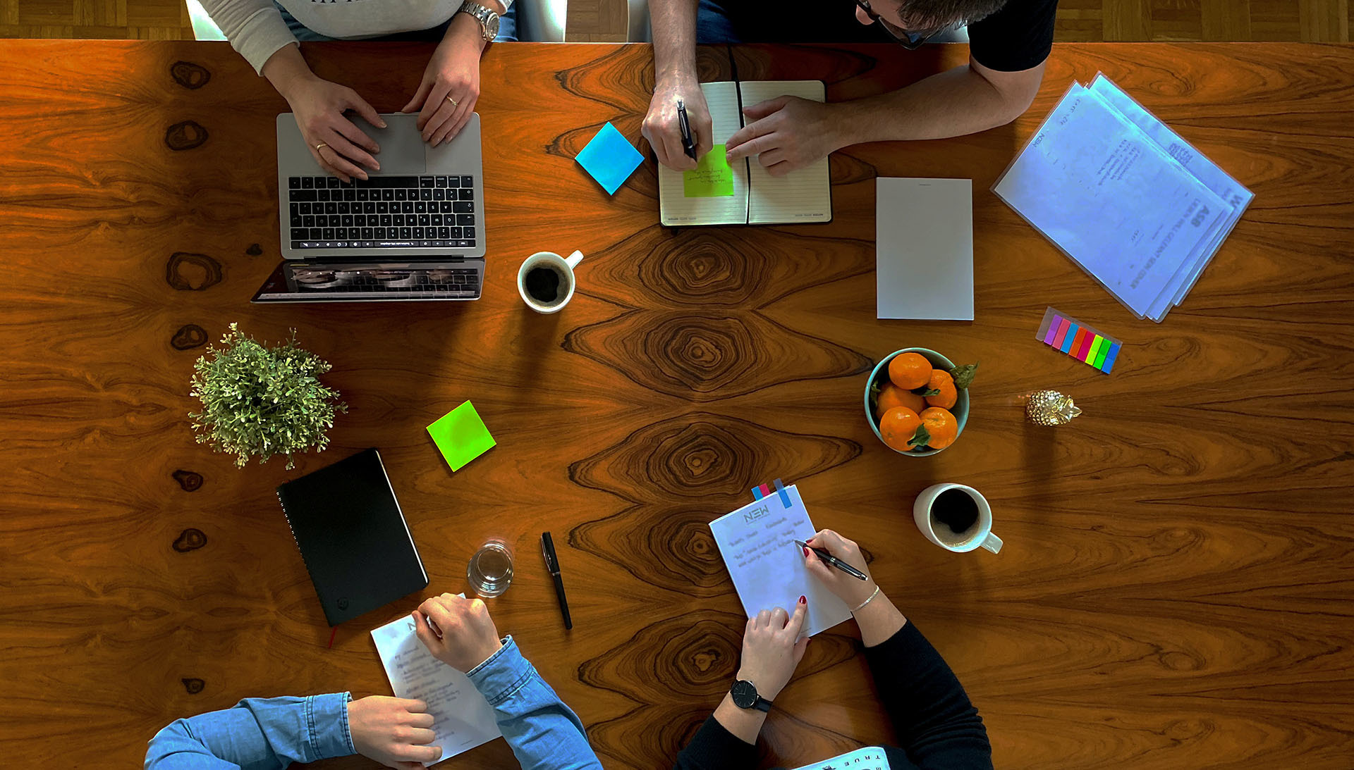 Kreativteam von N3W bei der Projektarbeit: Laptop, Notizbücher, Kaffee und Obst auf einem Holztisch symbolisieren Zusammenarbeit und kreative Strategieentwicklung.
